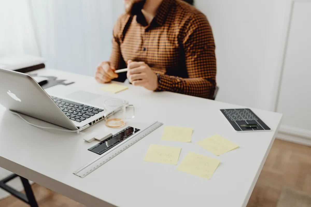 A person at a white desk with a laptop, ruler, notepad and handwritten sticky notes